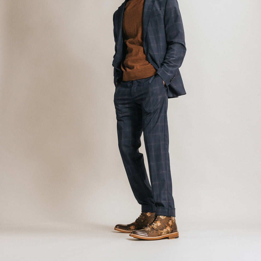A person dressed in a brown sweater, dark checkered suit, and ornate boots with hands in pockets against a plain background.