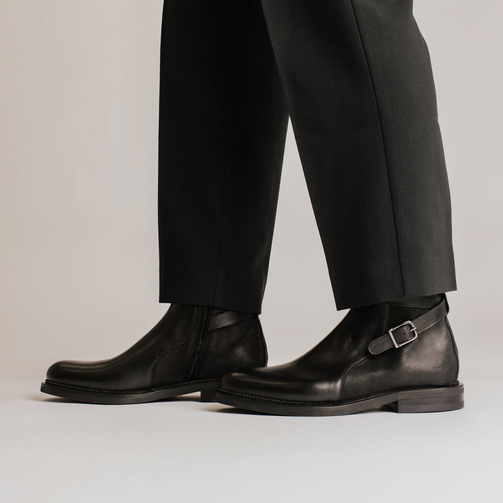 Person wearing black boots with silver buckle and black dress pants on a white background.