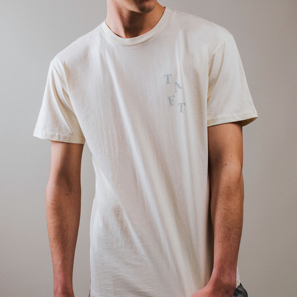 A person wearing a beige T-shirt stands against a plain background. The letters TAFT are printed on the left chest of the shirt.