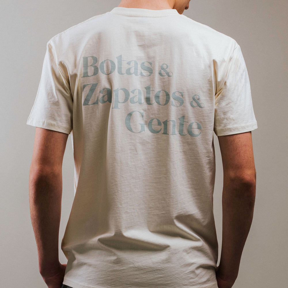 A person wearing a beige T-shirt with the text Botas & Zapatos & Gente printed on the back in grayish blue letters. The person is facing away from the camera.