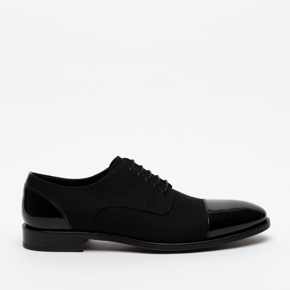 A single black oxford shoe with a polished cap toe and heel. The shoe has black laces and a low heel. The background is plain white.