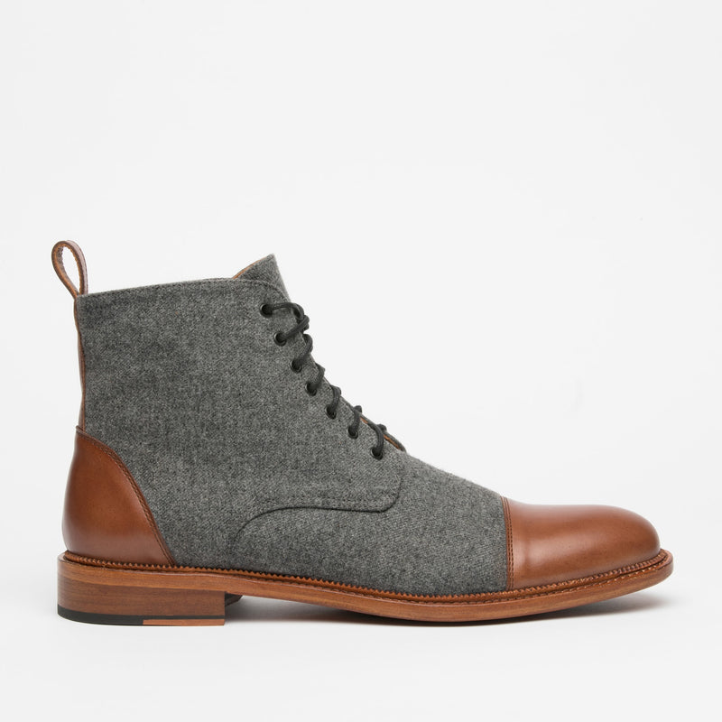A side view of a men's ankle boot with a gray wool upper, brown leather toe cap and heel, black laces, and a wooden sole.