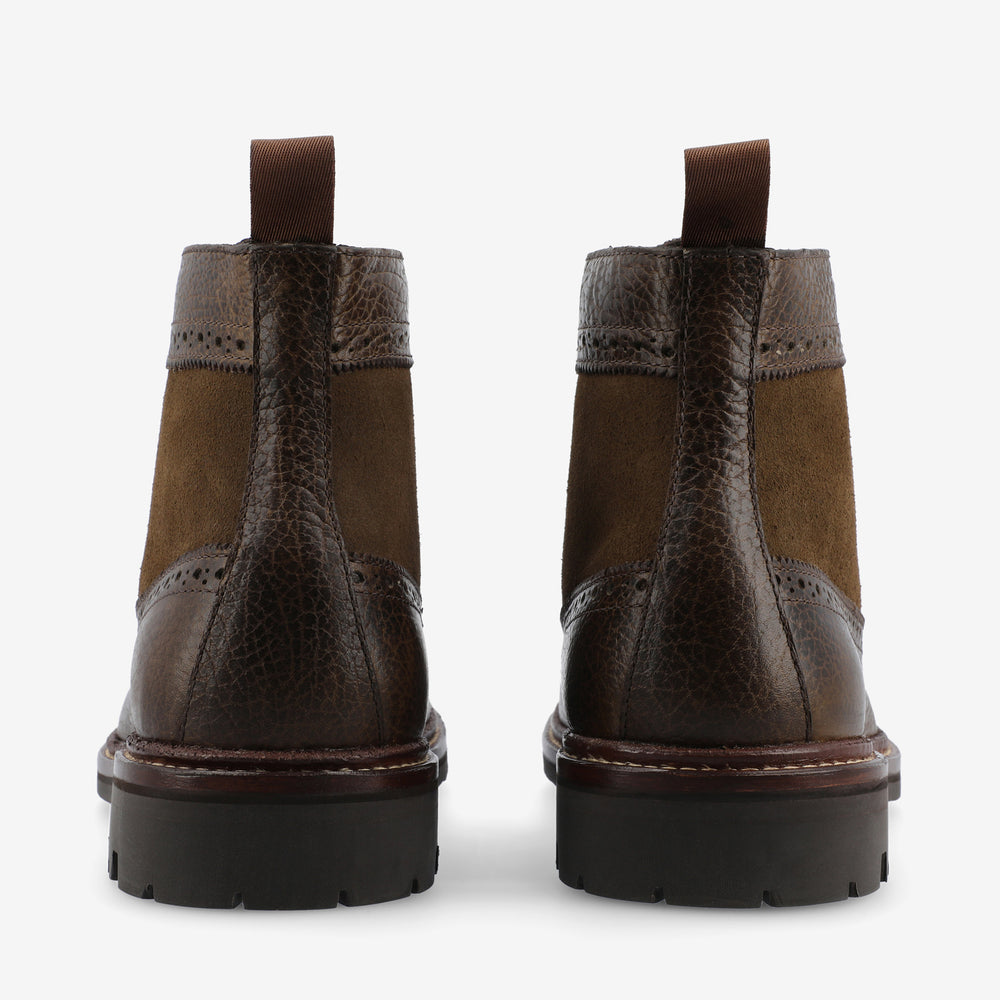 A pair of brown leather and suede boots are shown from the back, featuring a pull-tab on the heel and a rugged sole.