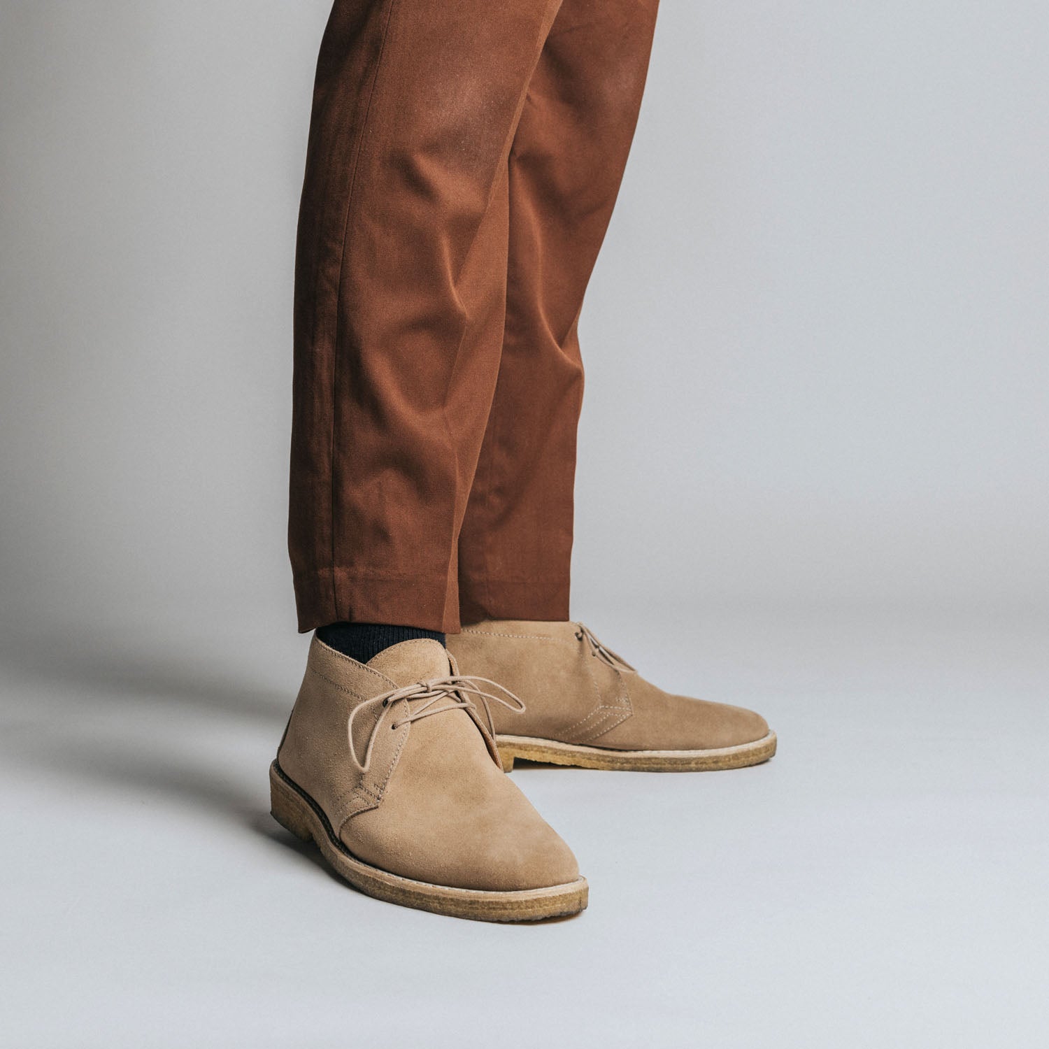 A person wearing brown suede desert boots and rust-colored pants stands against a plain gray background.