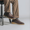 A person wearing brown leather shoes and khaki pants sits on a metal stool against a gray background.