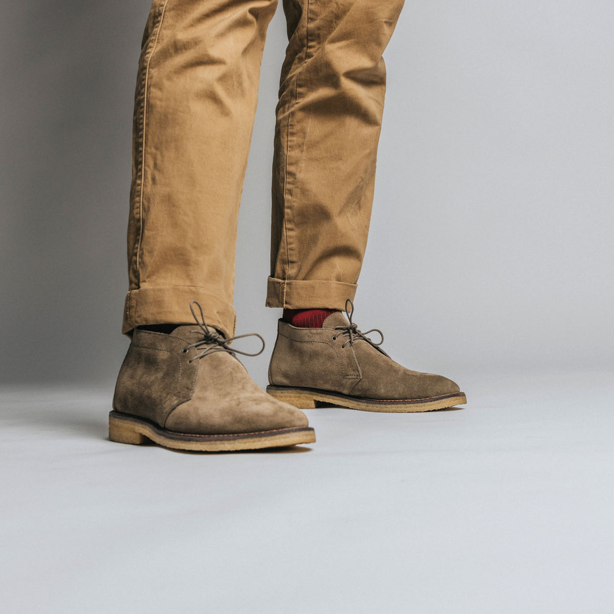 Person wearing light brown chinos and grey suede shoes with red socks, standing against a plain background.