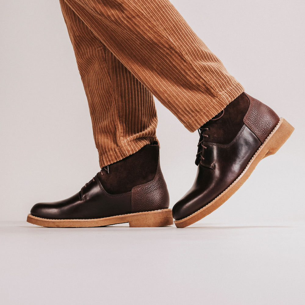 A person wearing brown corduroy pants and dark brown leather ankle boots with rubber soles walks against a neutral background.