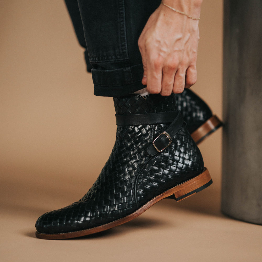 A person wearing black woven leather boots adjusts the strap while dressed in dark rolled-up pants.