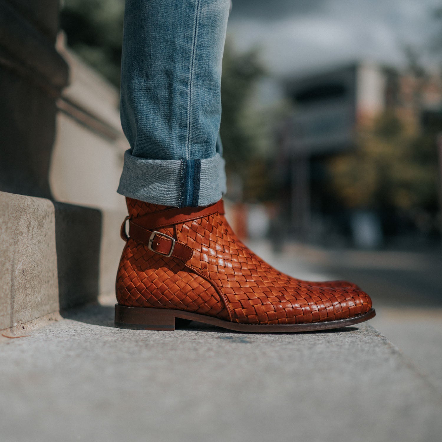 Close-up of a person wearing rolled-up jeans and woven brown leather ankle boots, standing on a concrete surface outdoors.