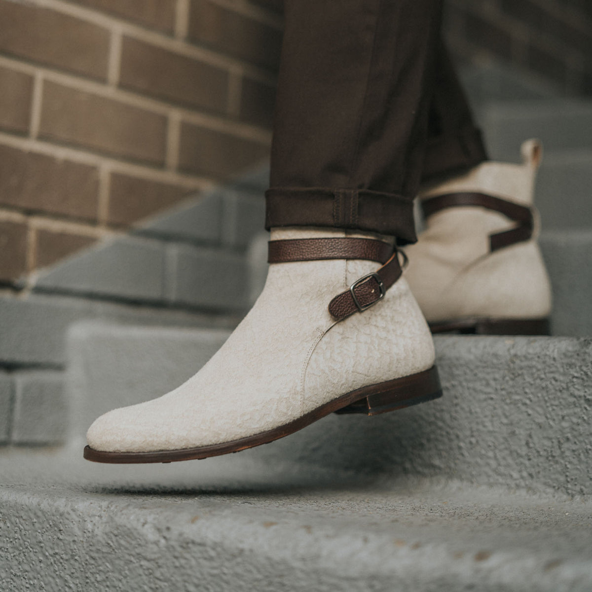 Person wearing beige suede ankle boots with brown straps, stepping on gray concrete stairs next to a brick wall.