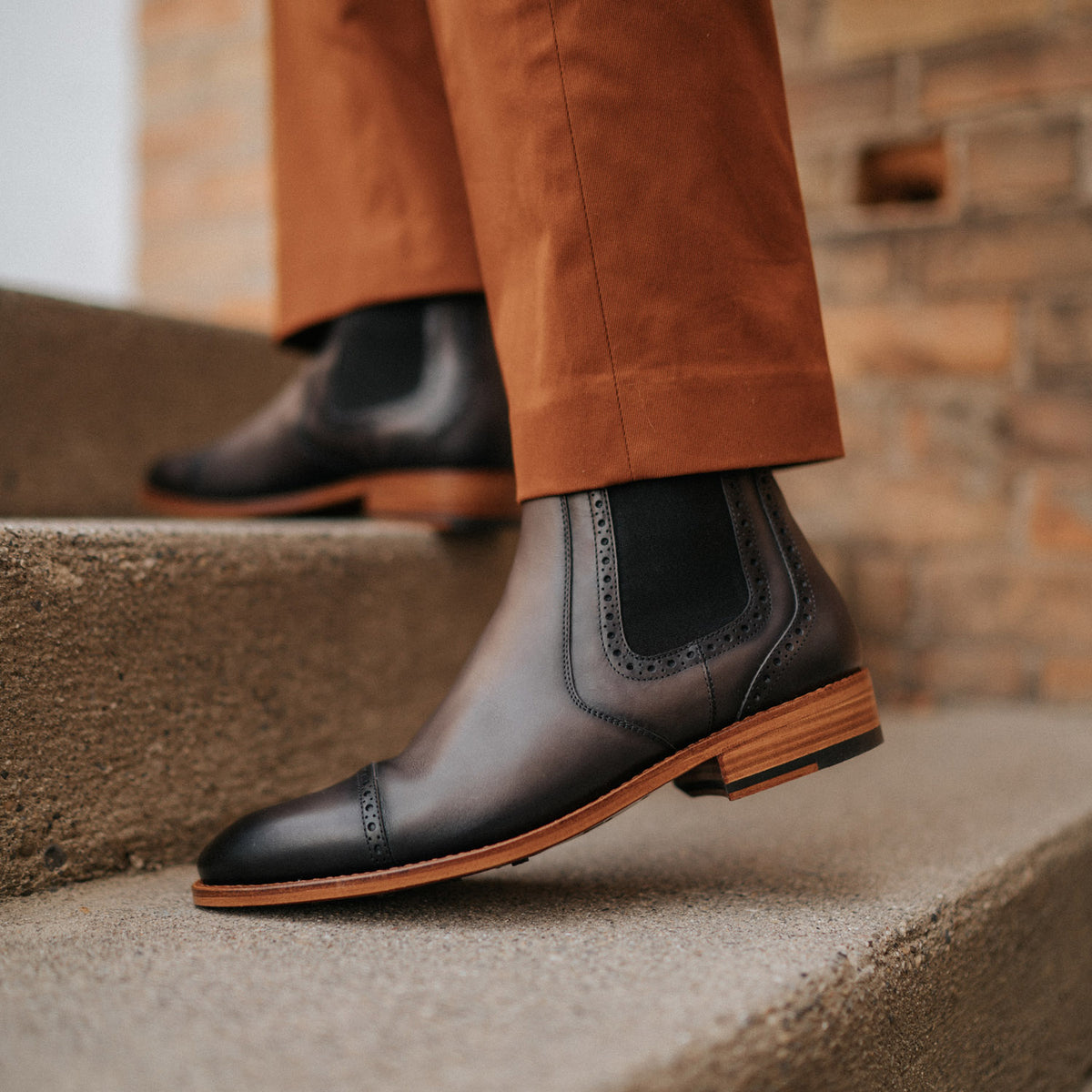 A person wearing brown pants is walking up concrete stairs in black leather ankle boots with subtle brogue detailing and wooden soles.