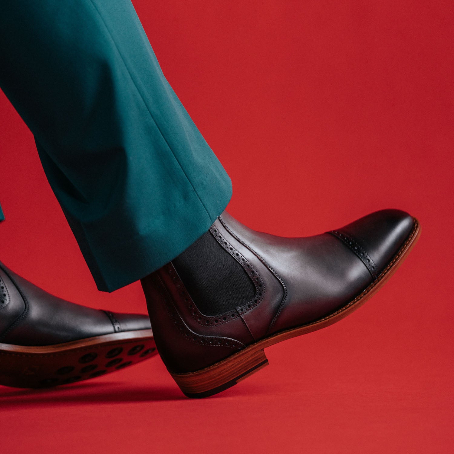 Close-up of a person wearing green pants and black leather dress boots with brogue detailing against a red background.