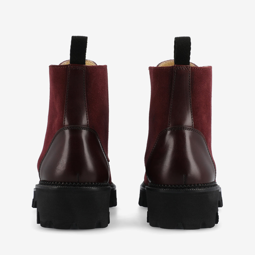 A pair of burgundy boots with chunky black soles, featuring a mix of suede and leather materials, viewed from the back.