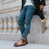 A person stands against a marble railing, showcasing a casual outfit with blue jeans and tan leather Chelsea boots.