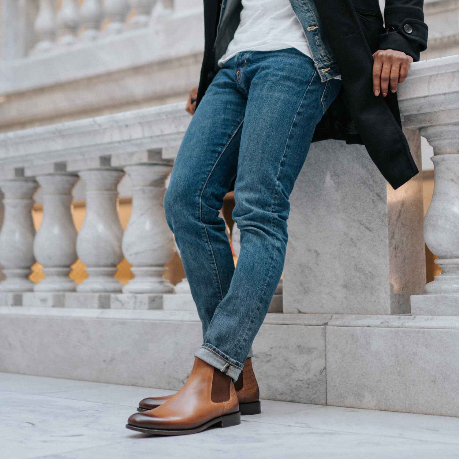 A person stands against a marble railing, showcasing a casual outfit with blue jeans and tan leather Chelsea boots.