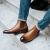 Two legs with cuffed jeans are shown wearing brown leather Chelsea boots with a textured panel, standing on marble steps.