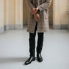 A person stands indoors wearing a checkered overcoat, black pants, and black boots, with their arms crossed and only the lower half of their body visible.