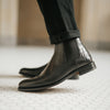 Close-up of a person wearing black leather ankle boots with a textured design on the back panel, paired with black jeans. The background shows a tiled floor.