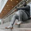 A man with a beard sits on outdoor stairs in front of a building, wearing grey pants, a dark jacket, and light-colored boots with brown accents.