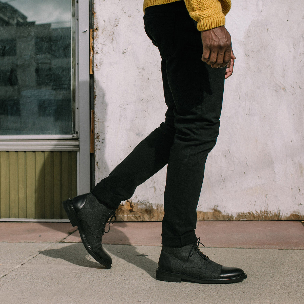 A person wearing a yellow sweater, black pants, and black-gray lace-up boots walks on a sidewalk next to a tall window and weathered wall.