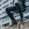 Person sitting on a concrete wall wearing floral patterned black boots, black jeans, and a black jacket in front of a modern glass building.