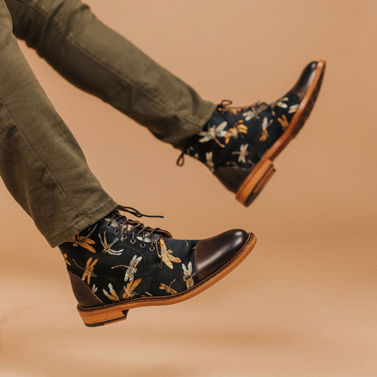 Person wearing dark leather boots with dragonfly designs, paired with olive green pants, against a beige background.