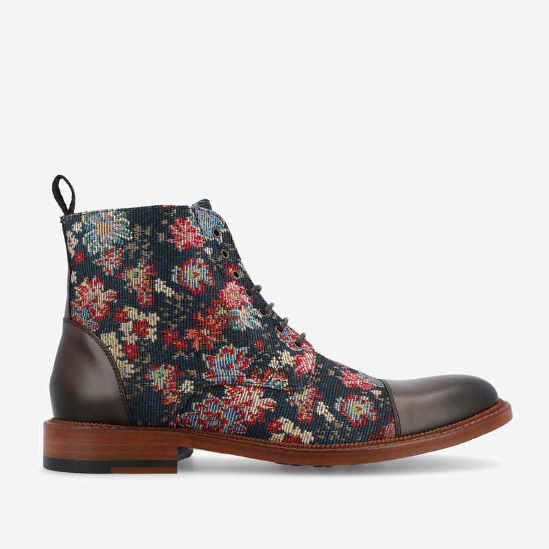 A side view of a lace-up ankle boot with a floral patterned fabric upper, dark brown leather toe and heel, and a wooden sole.