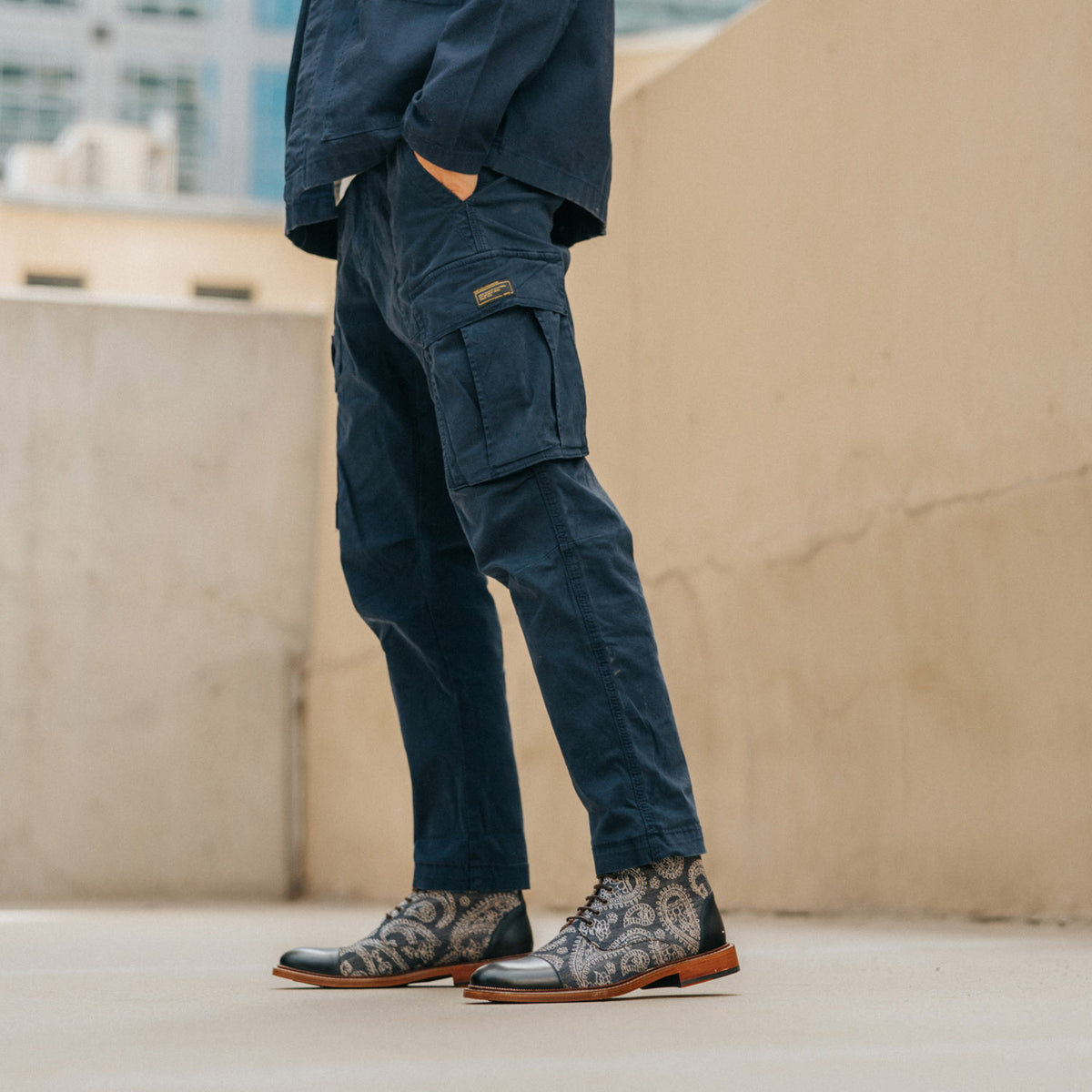 A person wearing navy cargo pants and paisley-patterned ankle boots stands on a concrete surface next to a beige wall in an outdoor setting.