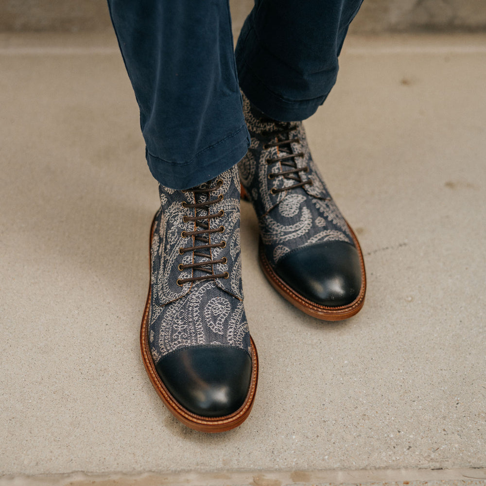 A person wearing blue trousers is shown from the knees down, highlighting their navy blue paisley-patterned lace-up shoes with brown soles.