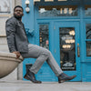 A man sits on a concrete structure in front of a blue door, wearing a gray jacket, gray pants, and patterned boots.