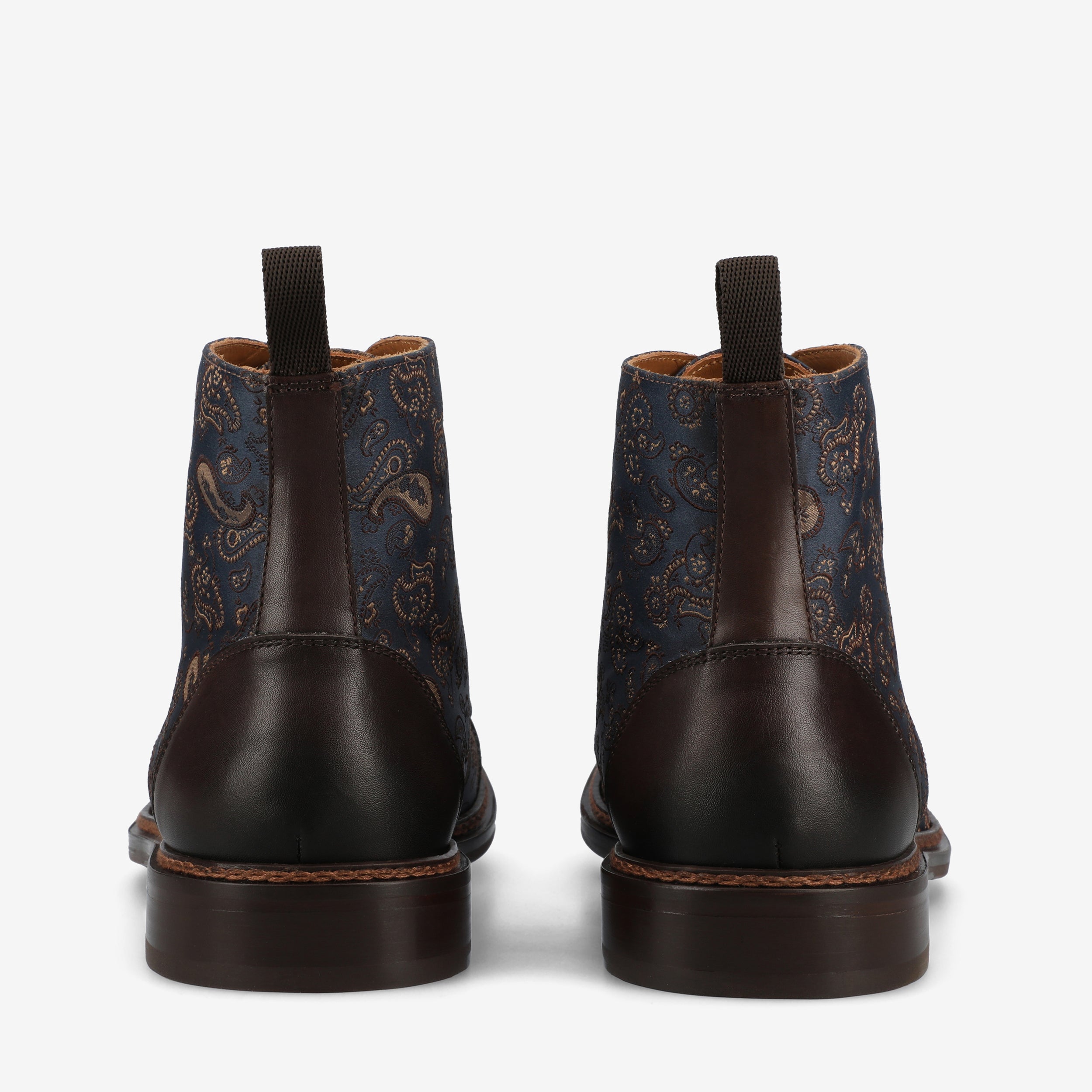 Rear view of a pair of ankle boots featuring dark brown leather on the lower half and navy blue fabric with a paisley pattern on the upper half. The boots have pull tabs at the back.