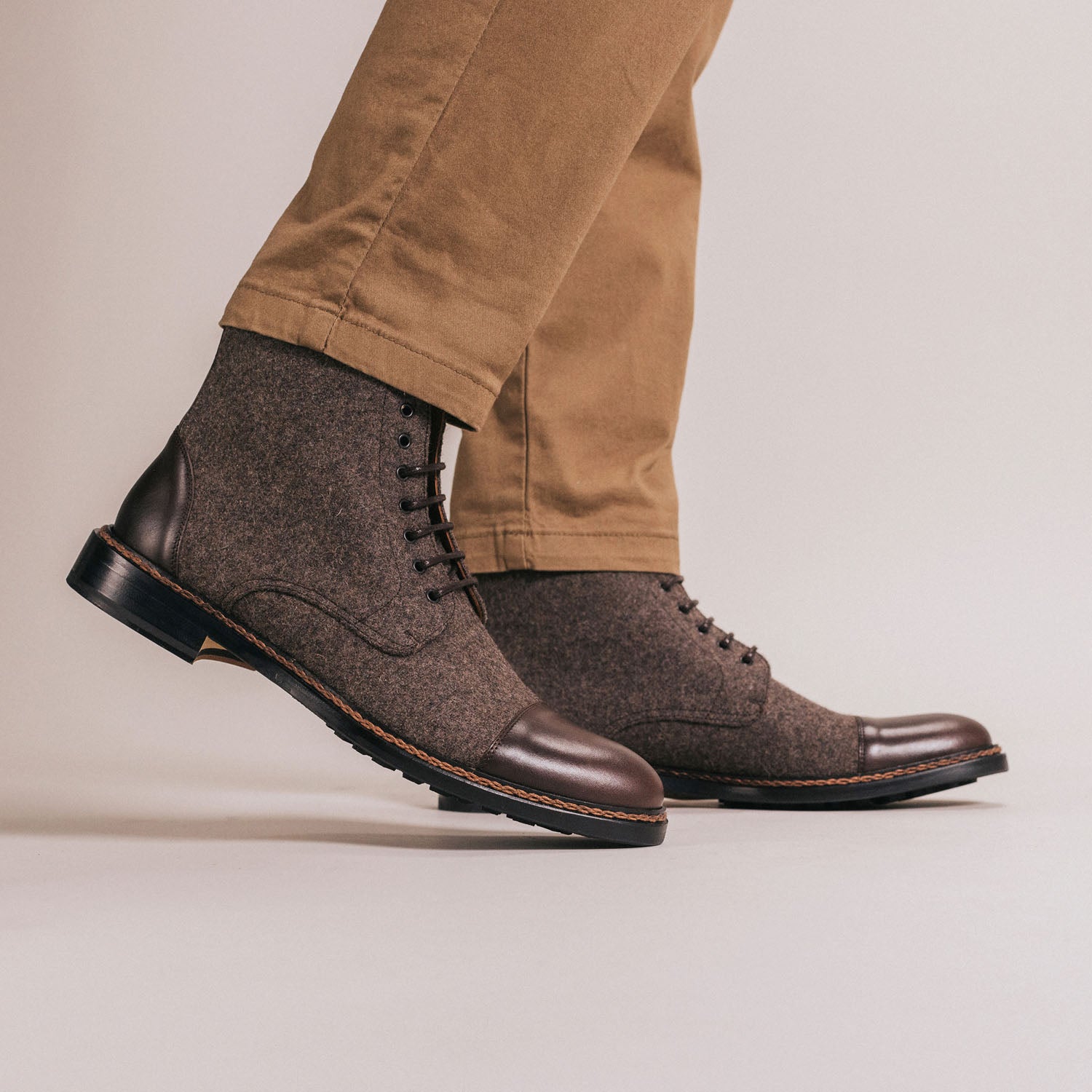 Person wearing beige pants and brown leather and wool ankle boots with black laces, standing on a neutral background.