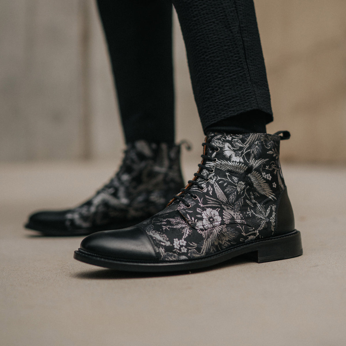 Close-up of person wearing black floral-patterned ankle boots with black pants, standing indoors on a light-colored surface.