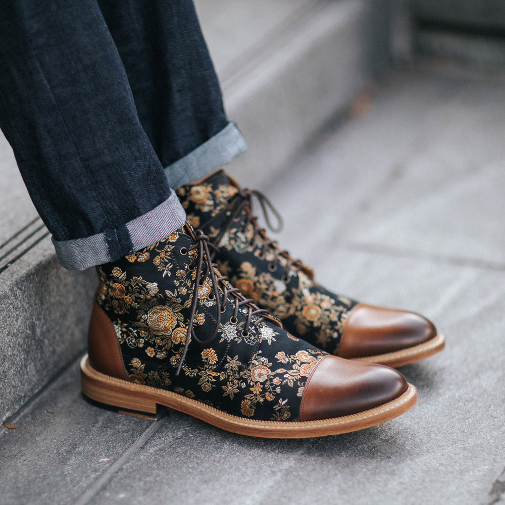 A person wearing dark jeans and floral-patterned brown leather lace-up boots stands on gray steps.