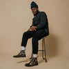 A man wearing a black beanie, black puffer jacket, dark pants, white socks, and floral boots sits on a stool against a beige background.