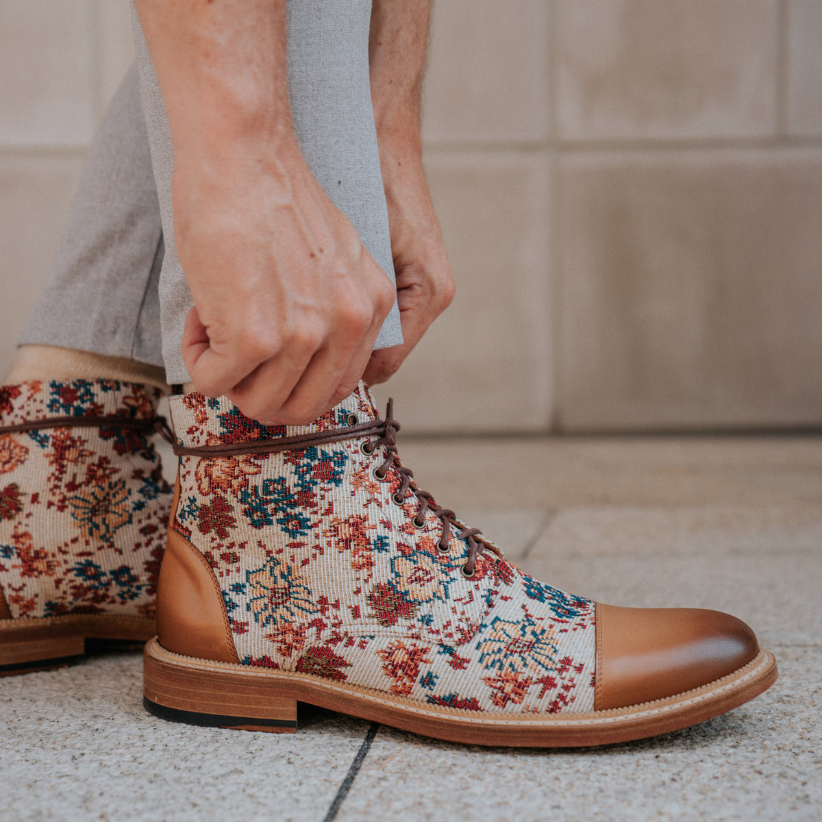 Close-up of a person tying the laces of a floral-patterned boot with brown leather accents. The person is wearing light gray pants, and the ground appears to be a tiled floor.