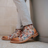 Person wearing light gray pants and floral patterned boots with brown leather toe caps and heels stands next to a wall.