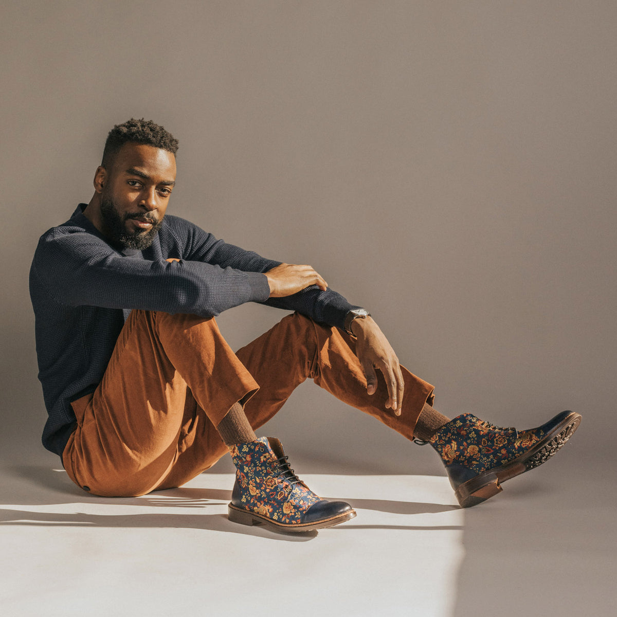 A man with a beard sits on the floor against a neutral background, wearing a navy sweater, brown pants, and blue floral boots.