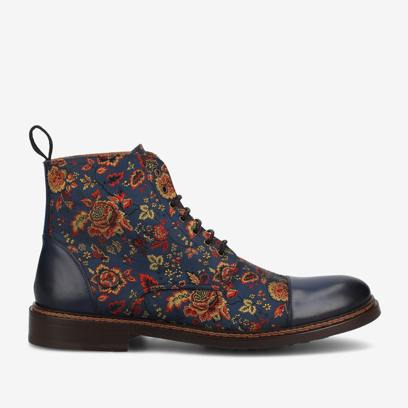 A side view of a lace-up boot with dark blue leather at the toe and heel, featuring a floral fabric design in red, yellow, and green on a dark blue background.