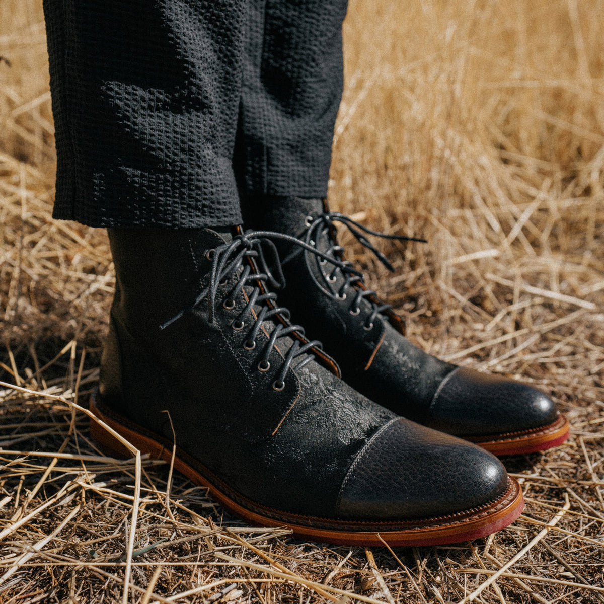 Person wearing black leather lace-up boots stands on dry, grassy ground. Only the lower legs and boots are visible, with the individual dressed in dark textured pants.