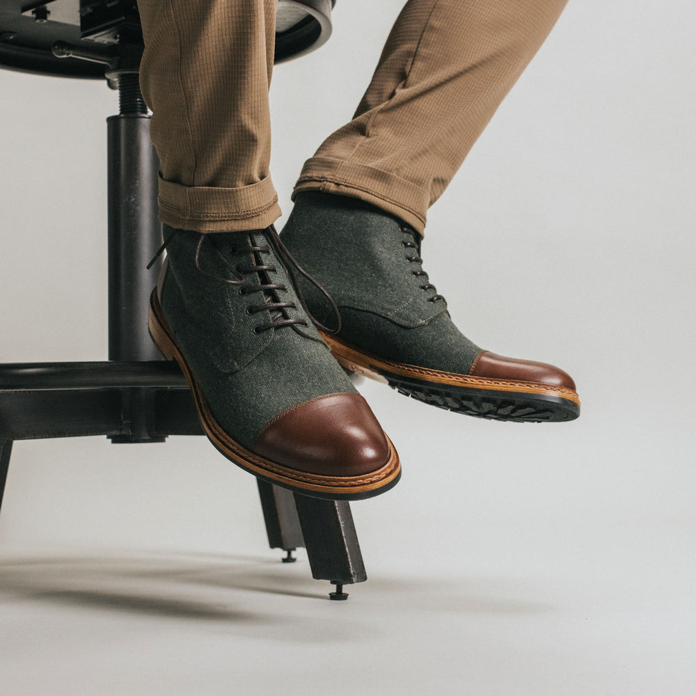 A person wearing gray and brown leather boots sits on a black stool. Their legs are clad in light brown pants.