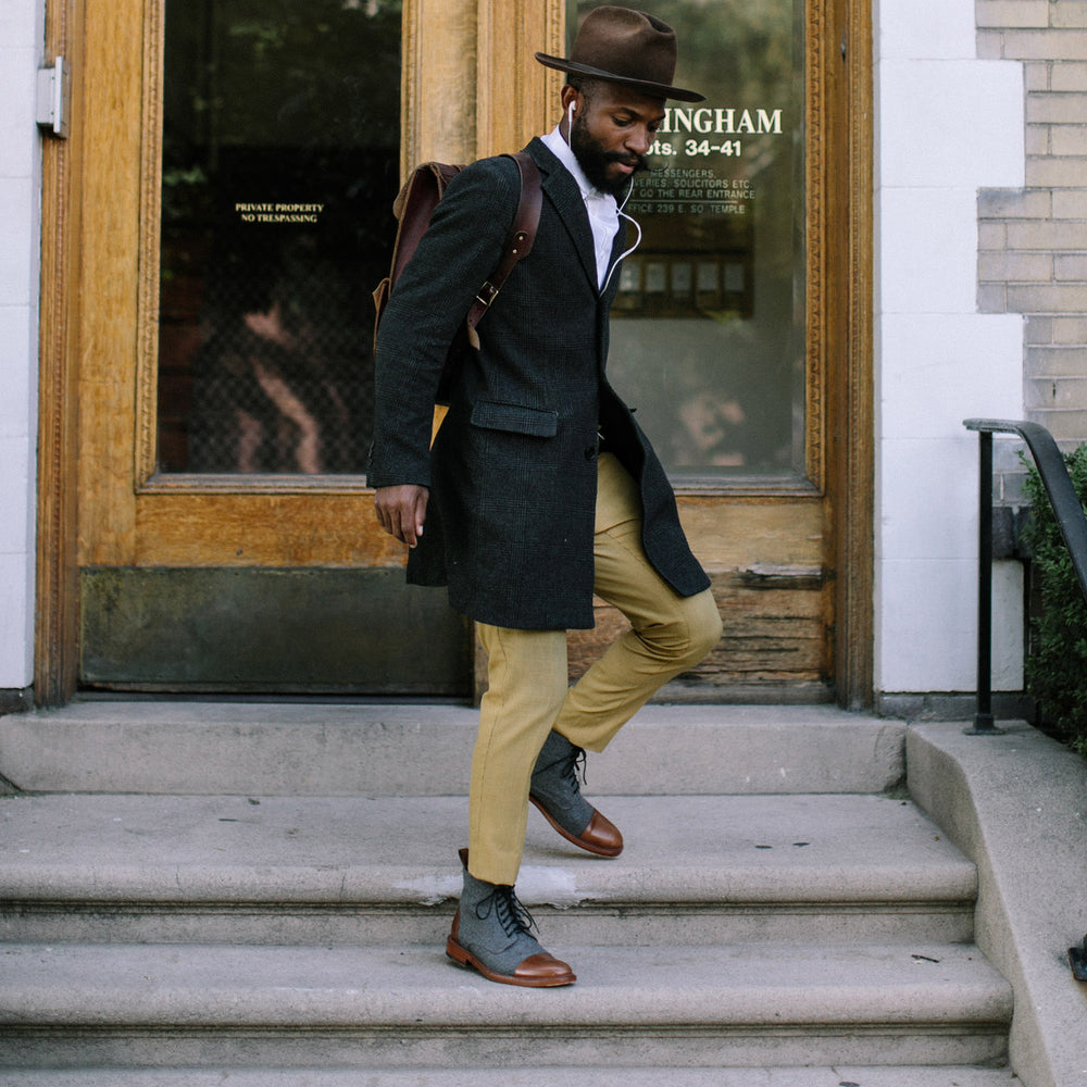 A person in a dark coat, light pants, grey boots, and a brown hat walks down steps carrying a brown leather bag.