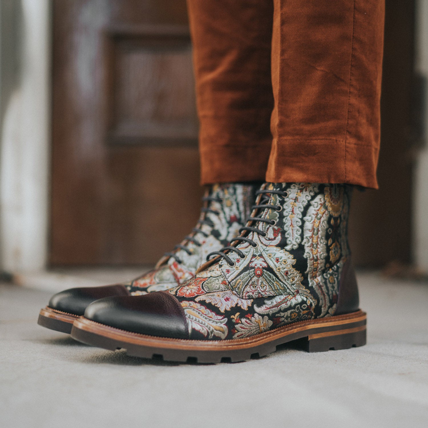 A person wearing detailed patterned boots with thick soles, paired with burnt orange trousers, standing on a light-colored surface in front of a wooden door.