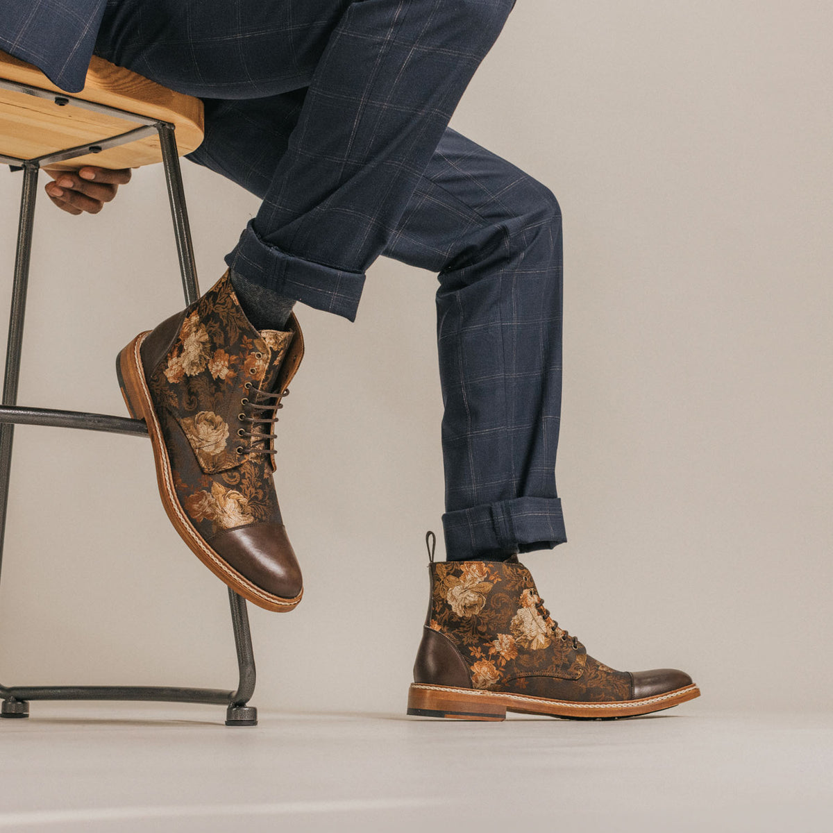 A person wearing blue plaid pants sits on a wooden stool, showcasing brown floral patterned boots with leather accents and a thick sole.