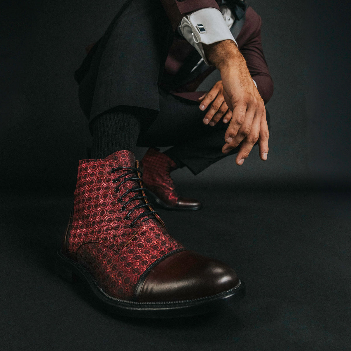 Man in dark suit crouching, showcasing red patterned dress boots with dark toes and laces.