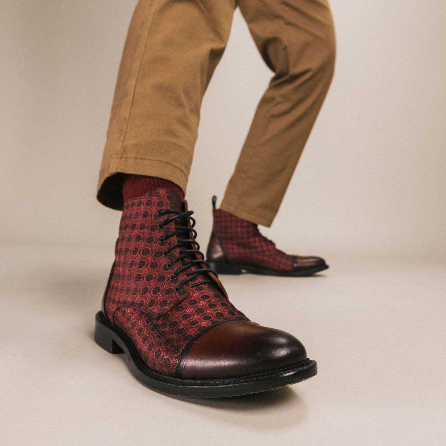 A person wearing brown pants and red patterned lace-up boots is shown from the waist down, with the focus on the footwear.