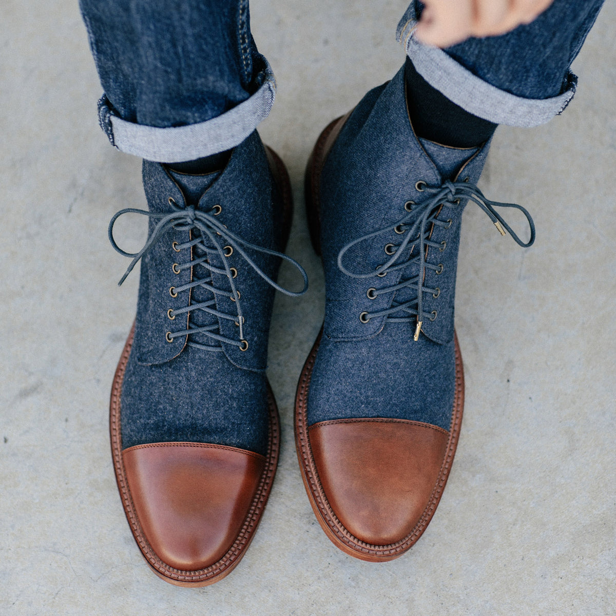 A pair of feet in two-tone boots with brown leather caps and dark material upper, worn with rolled-up jeans.