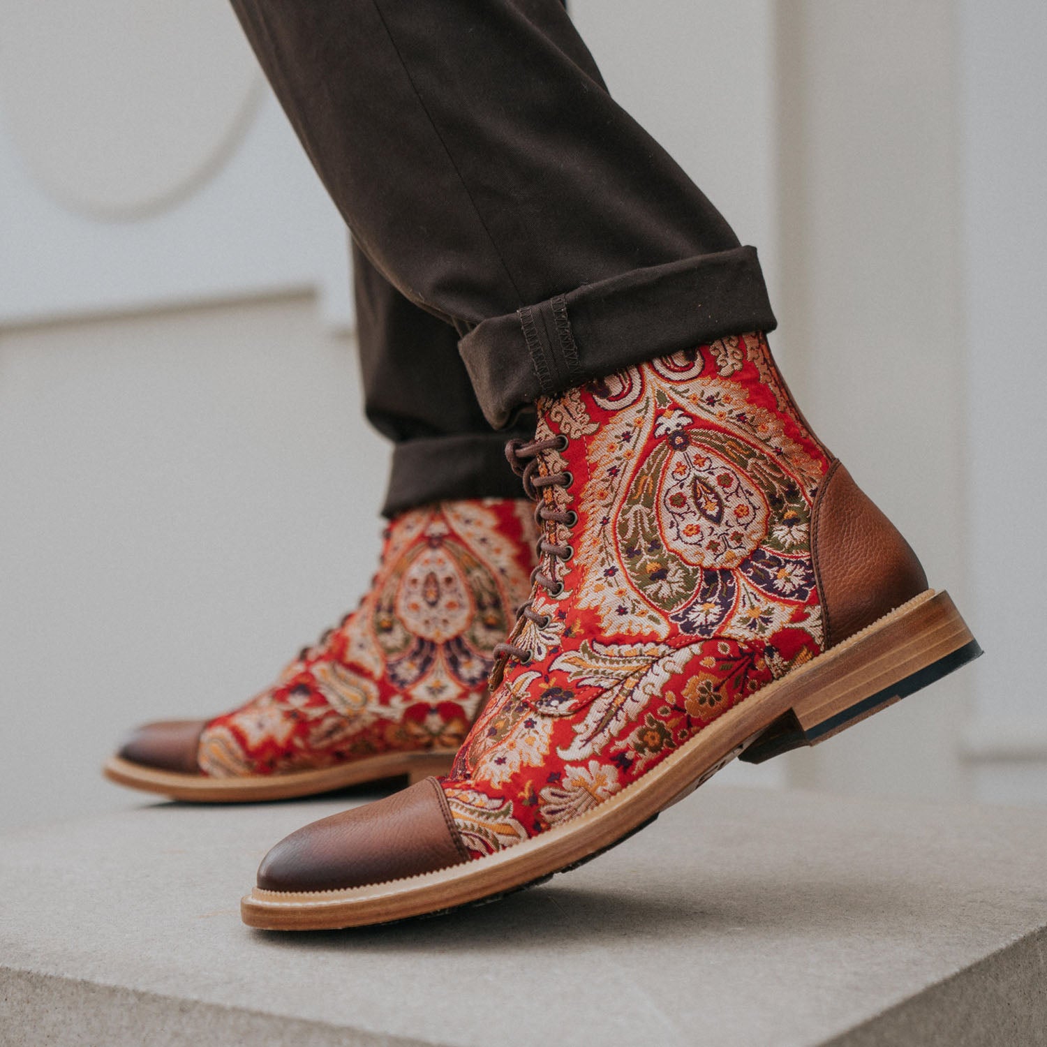 A person wearing paisley-patterned red and brown lace-up boots with dark pants, standing on a concrete surface.