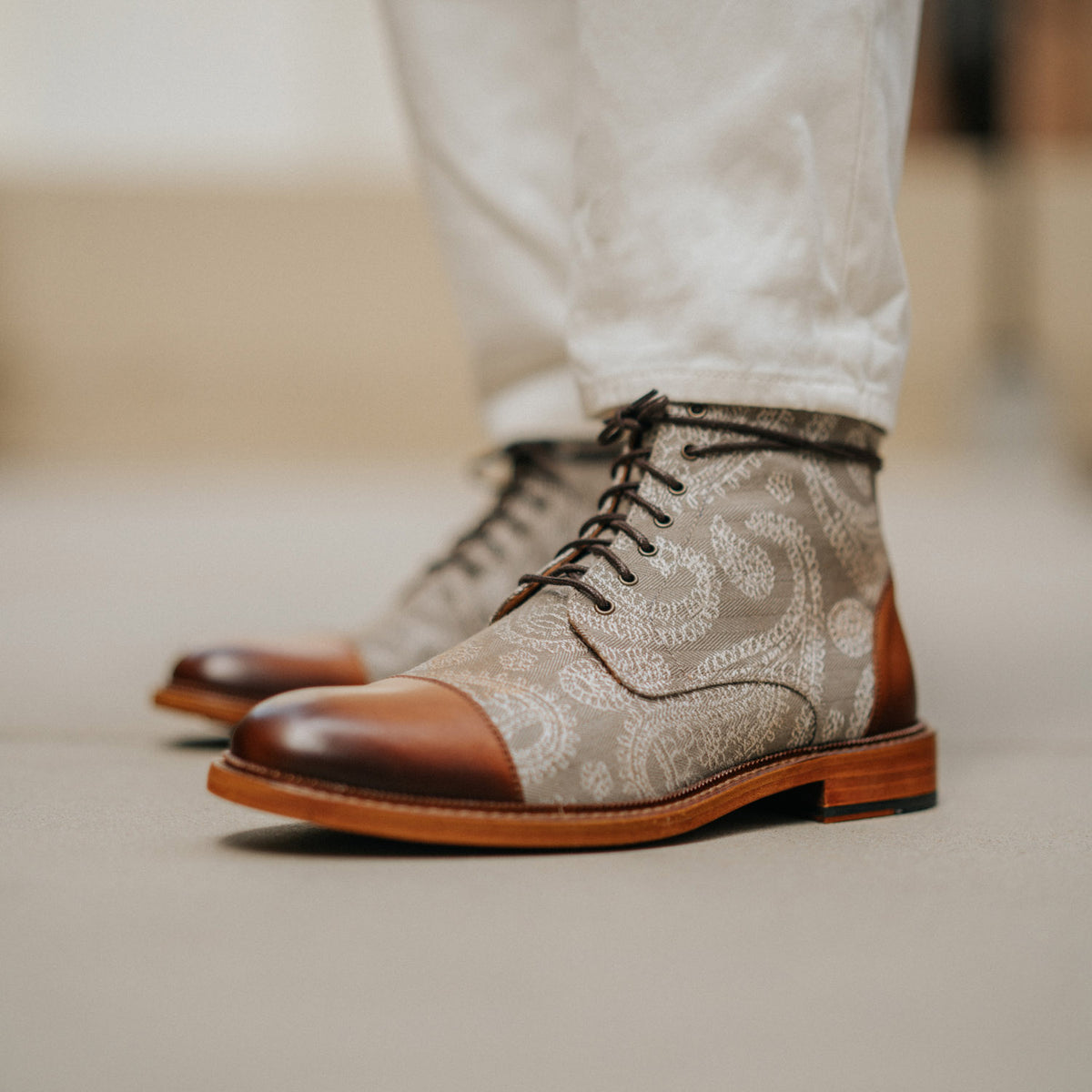 A pair of brown and white paisley-patterned lace-up boots worn by someone in white pants.