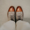 Top-down view of a person wearing brown leather and patterned beige fabric boots, with the tips polished to a sheen. The person is also wearing light-colored pants.